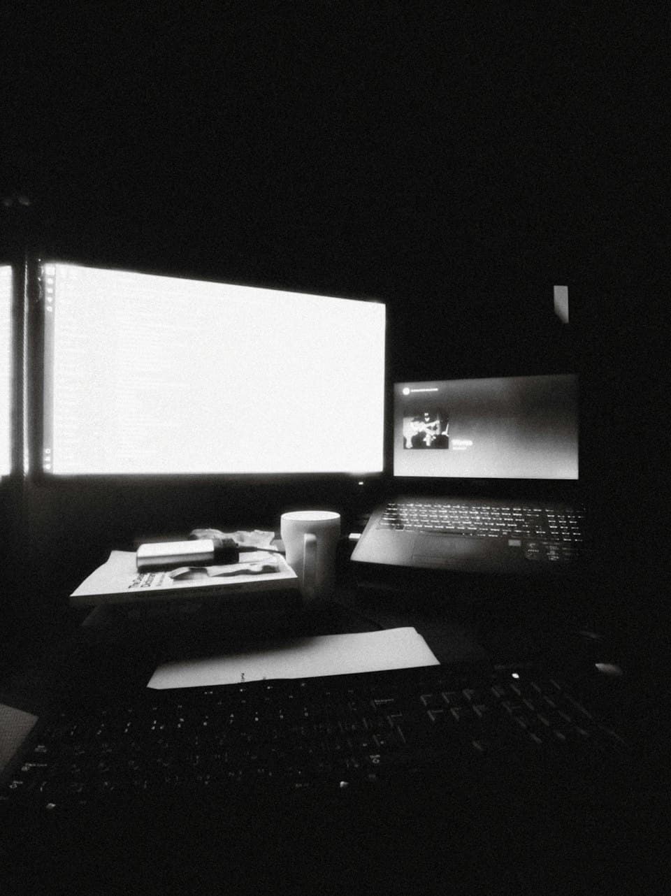 Schreibtisch mit leuchtenden Monitoren und Laptop in Schwarz-Weiß