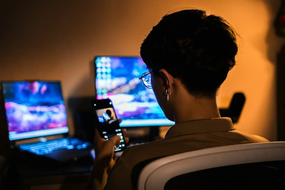 Person mit Smartphone vor leuchtenden Computerbildschirmen in einem dunklen Raum
